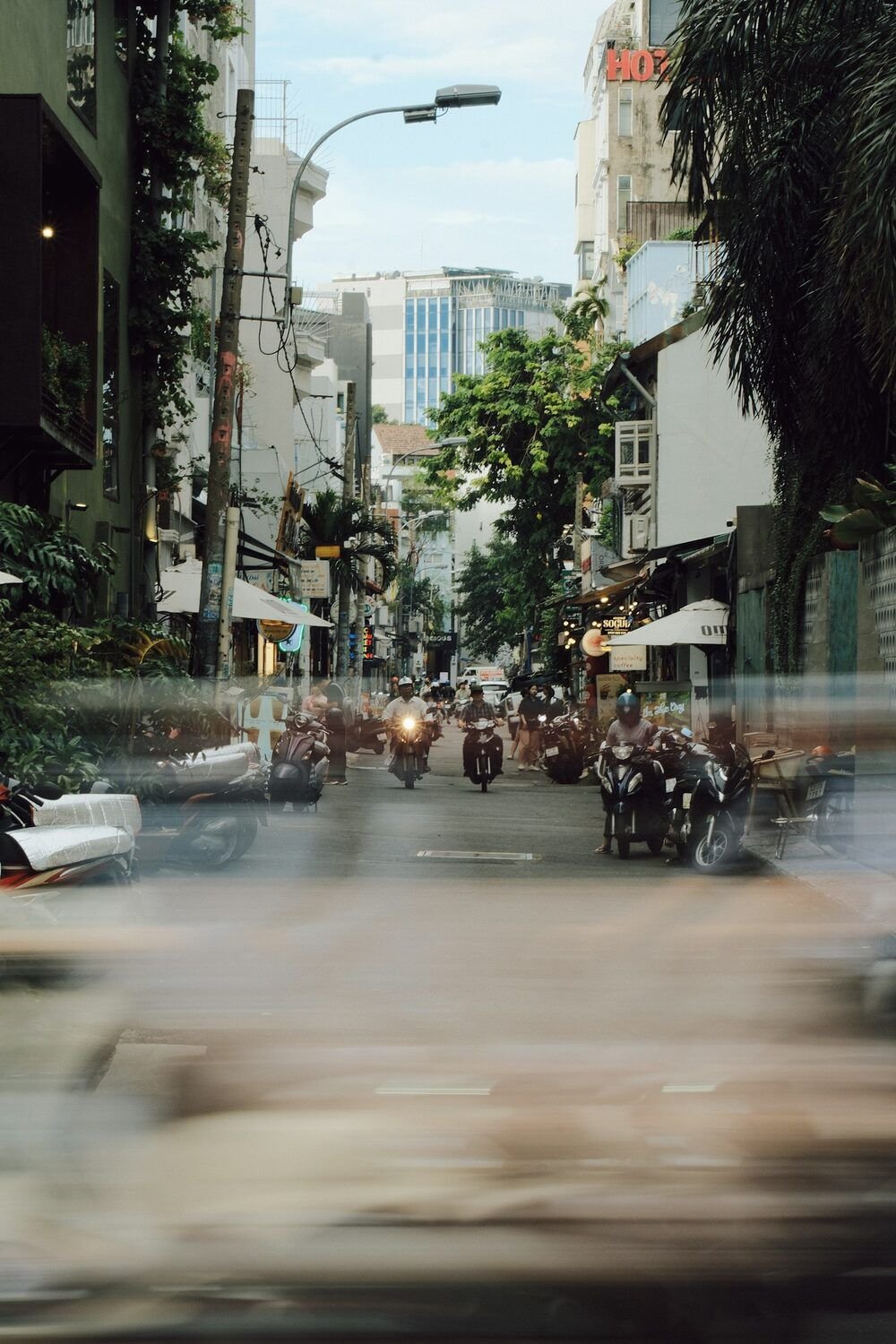 A tree-lined side street in Saigon with parked scooters and a few motorbikes riding towards the camera, the foreground smeared by motion blur