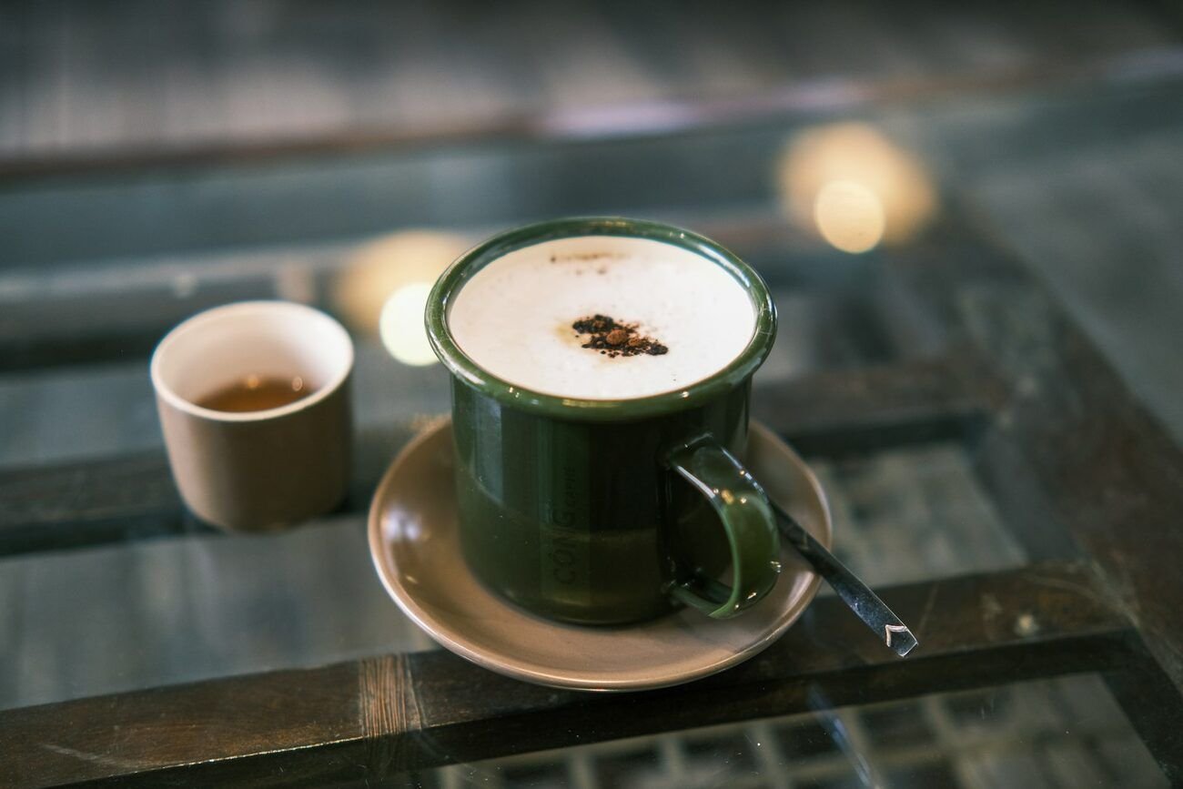 A cappuccino in a dark green enamel mug on a glass-topped table, with a small cup of tea beside it, in a Da Nang café